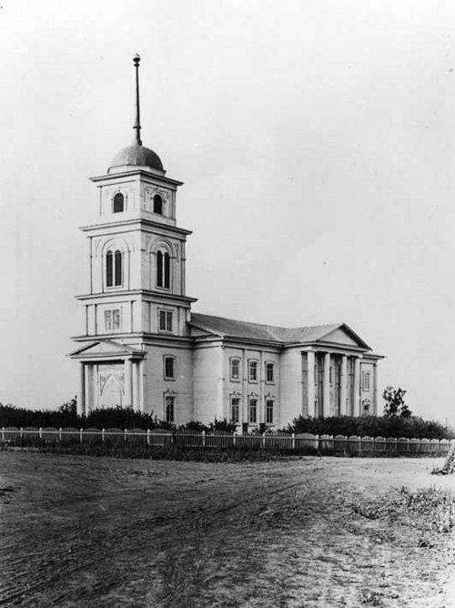 Die reformierte Kirche der Kolonie Norka mit Glockenturm und Säulenportal, davor ein Zaun und die unbefestigte Dorfstraße